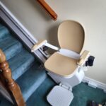 Cream covered stairlift with white base and frame. On a blue carpet covered staircase with cream walls